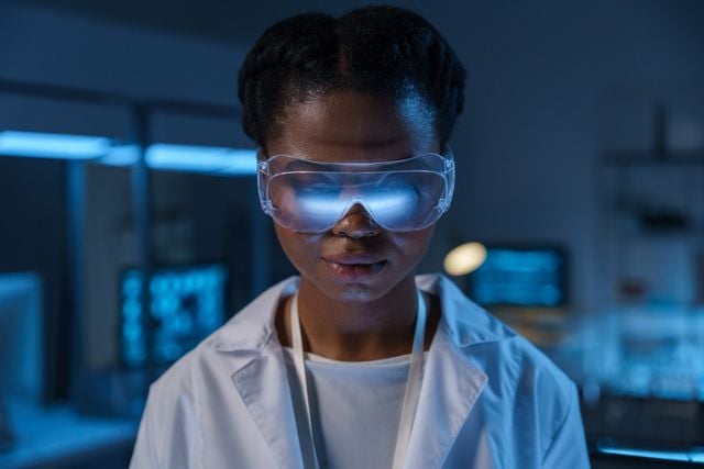 A medical laboratory researcher wearing safety goggles is illuminated by the blue glow from a monitor