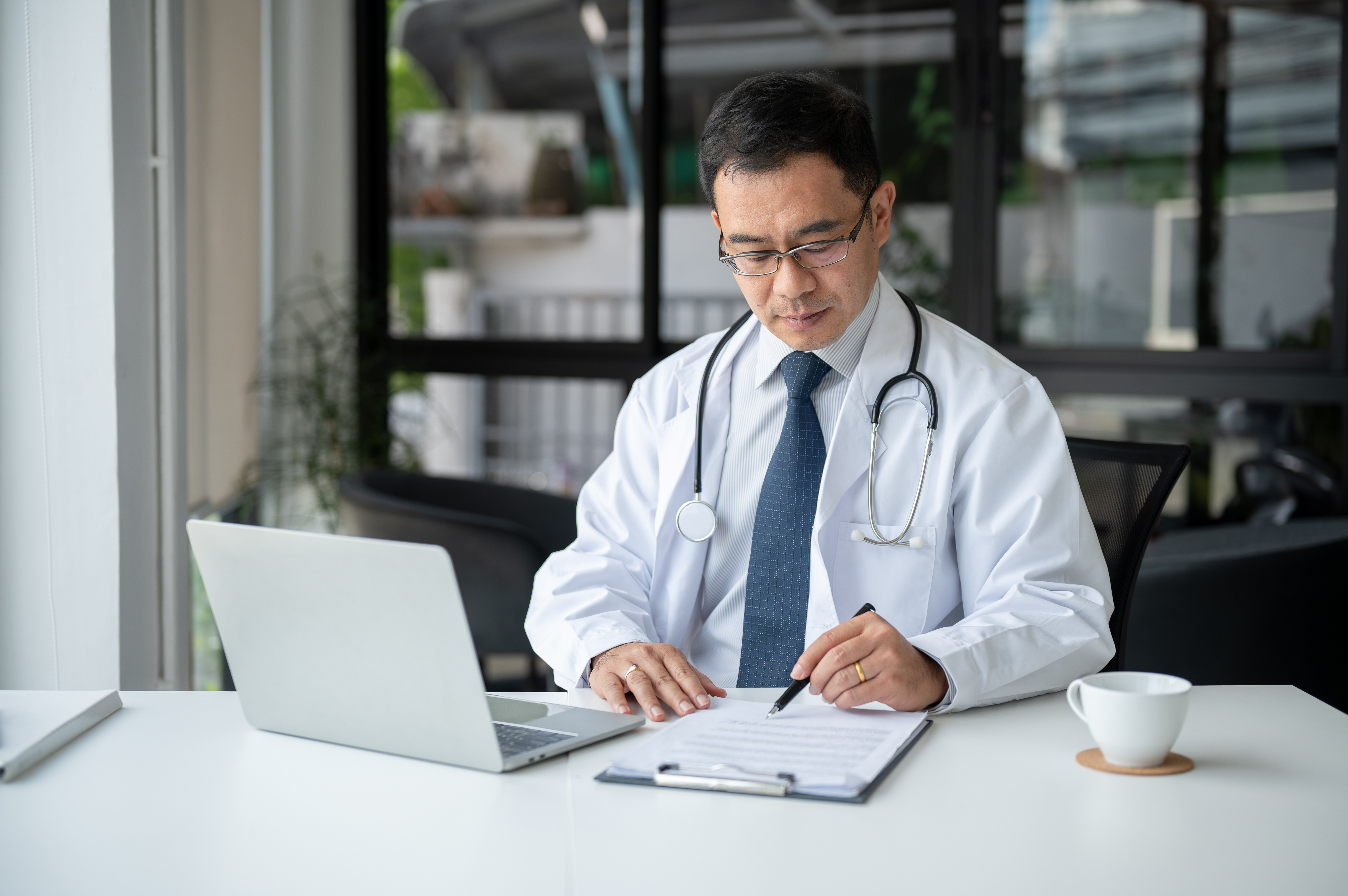 Physician writing a medical case report with clinical notes and a laptop.